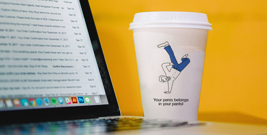A photograph of a paper coffee cup next to a computer, the coffee cup has a cartoon man doing a handstand and the slogan 'Your penis belongs in your pants!'